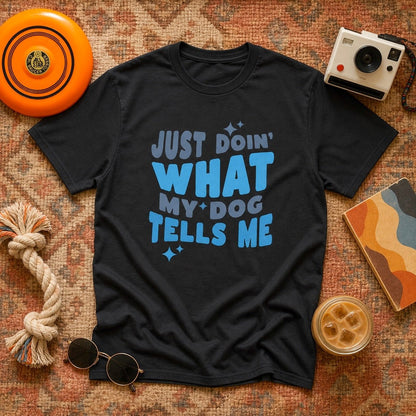 Black "Just Doin' What My Dog Tells Me" t-shirt in a flat lay with an orange frisbee, Polaroid camera, rope dog toy, round sunglasses, and iced coffee on a vintage patterned rug