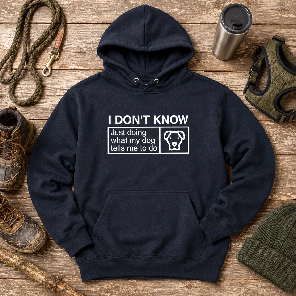 Navy pullover hoodie flat-lay with a white classification-label print reading "I DON'T KNOW — Just doing what my dog tells me to do" alongside a dog-face icon, styled with muddy hiking boots, a rope leash with brass clip, stainless steel travel tumbler, olive green dog harness, and ribbed knit beanie on weathered wood planks.