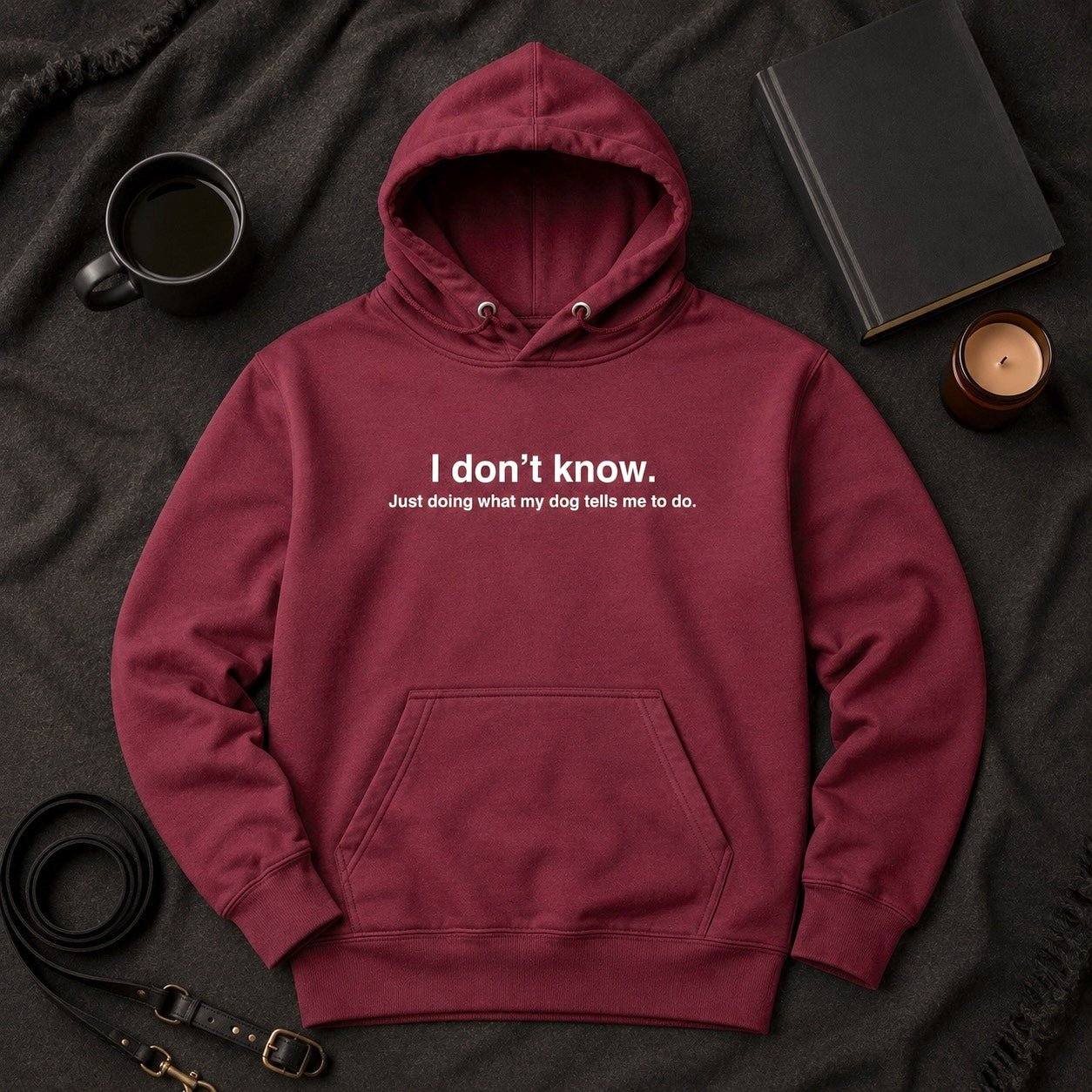 Maroon pullover hoodie flat-lay with white text reading "I don't know. Just doing what my dog tells me to do." styled with a black coffee mug, hardcover journal, amber candle, and black leather leash on a charcoal knit throw.