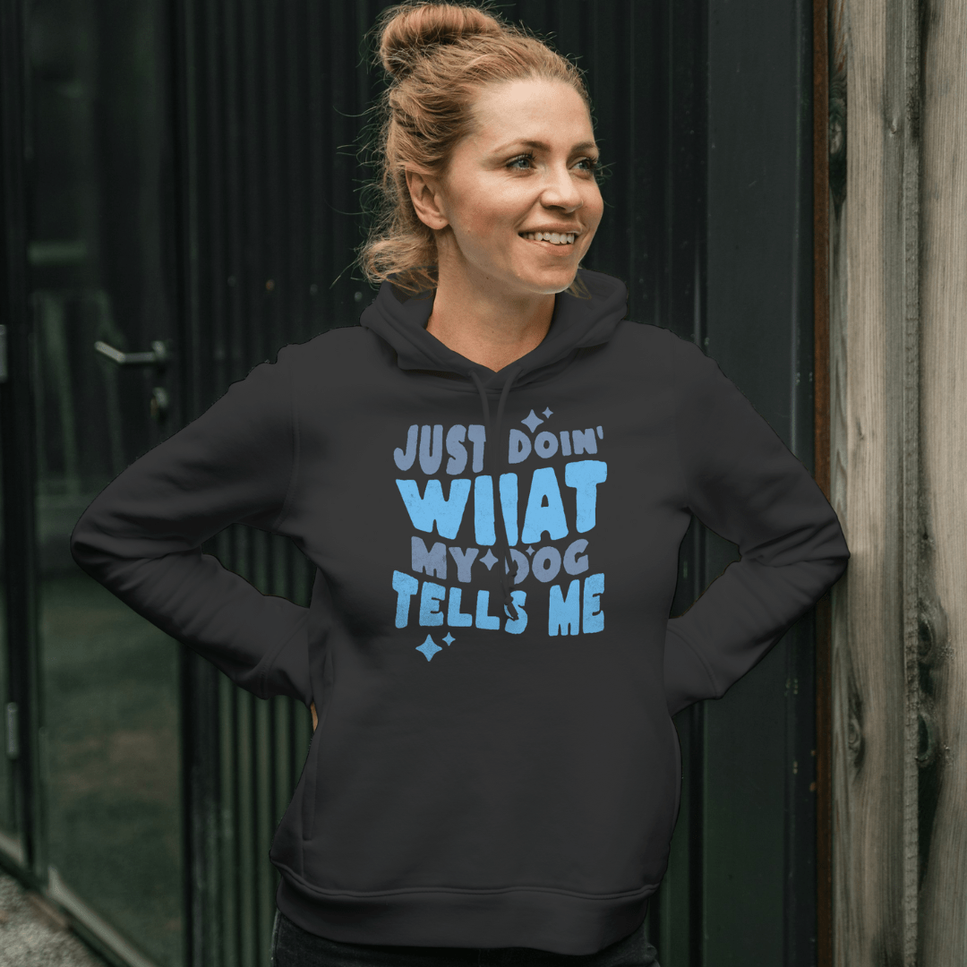 Against a dark metal wall, a person smiles, wearing the THiNK LiKE A DOG® black hoodie with "Just Doing What My Dog Tells Me" in blue. This cozy & fun dog lover apparel is perfect for expressing affection through fashion.