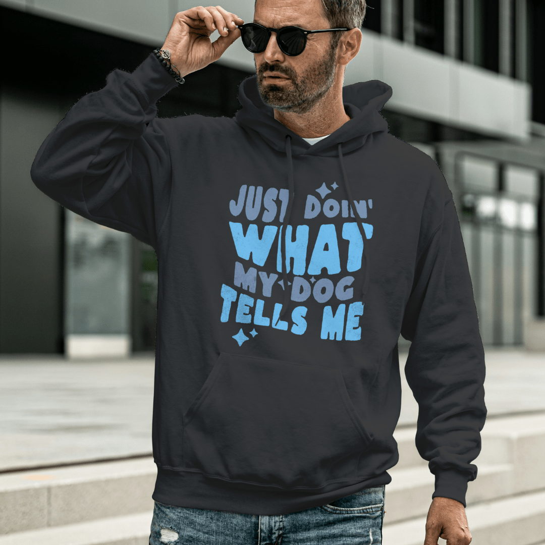 A man in sunglasses wears the "Just Doing What My Dog Tells Me" hoodie by THiNK LiKE A DOG®. This dark, dog-themed apparel is ideal for dog lovers and features playful text in blue, making it the perfect gift to celebrate their bond.