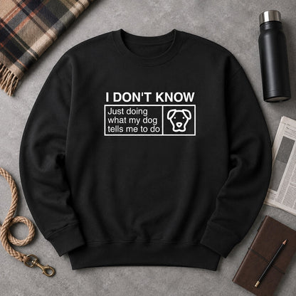 Black crewneck sweatshirt flat-lay with a white classification-label print reading "I DON'T KNOW — Just doing what my dog tells me to do" alongside a dog-face icon, styled with a plaid flannel throw, black insulated water bottle, folded newspaper, leather journal with pencil, and rope leash on slate concrete.