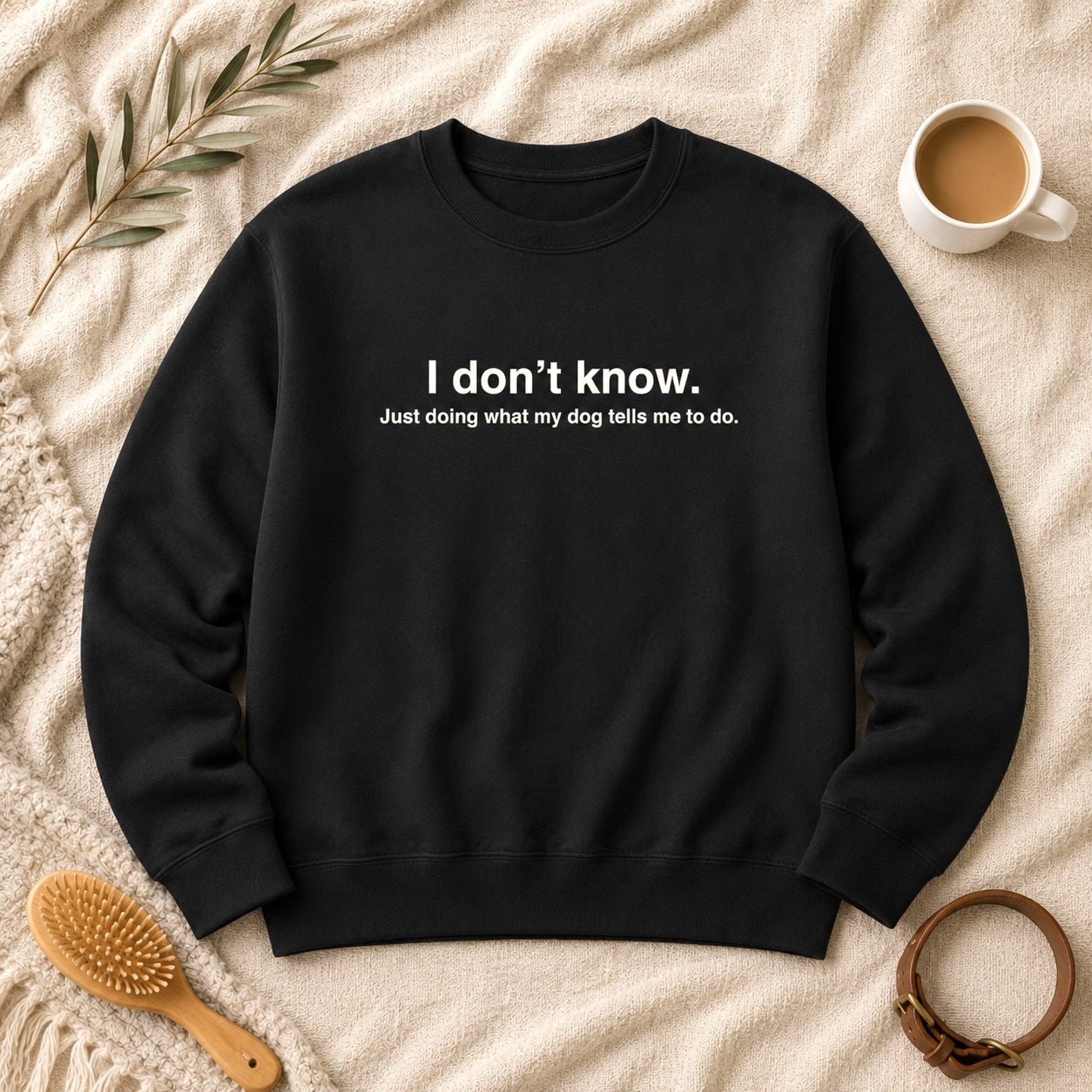 Black crewneck sweatshirt flat-lay with white text reading "I don't know. Just doing what my dog tells me to do." styled with an olive branch, latte mug, wooden grooming brush, leather dog collar, and chunky knit throw on cream linen.