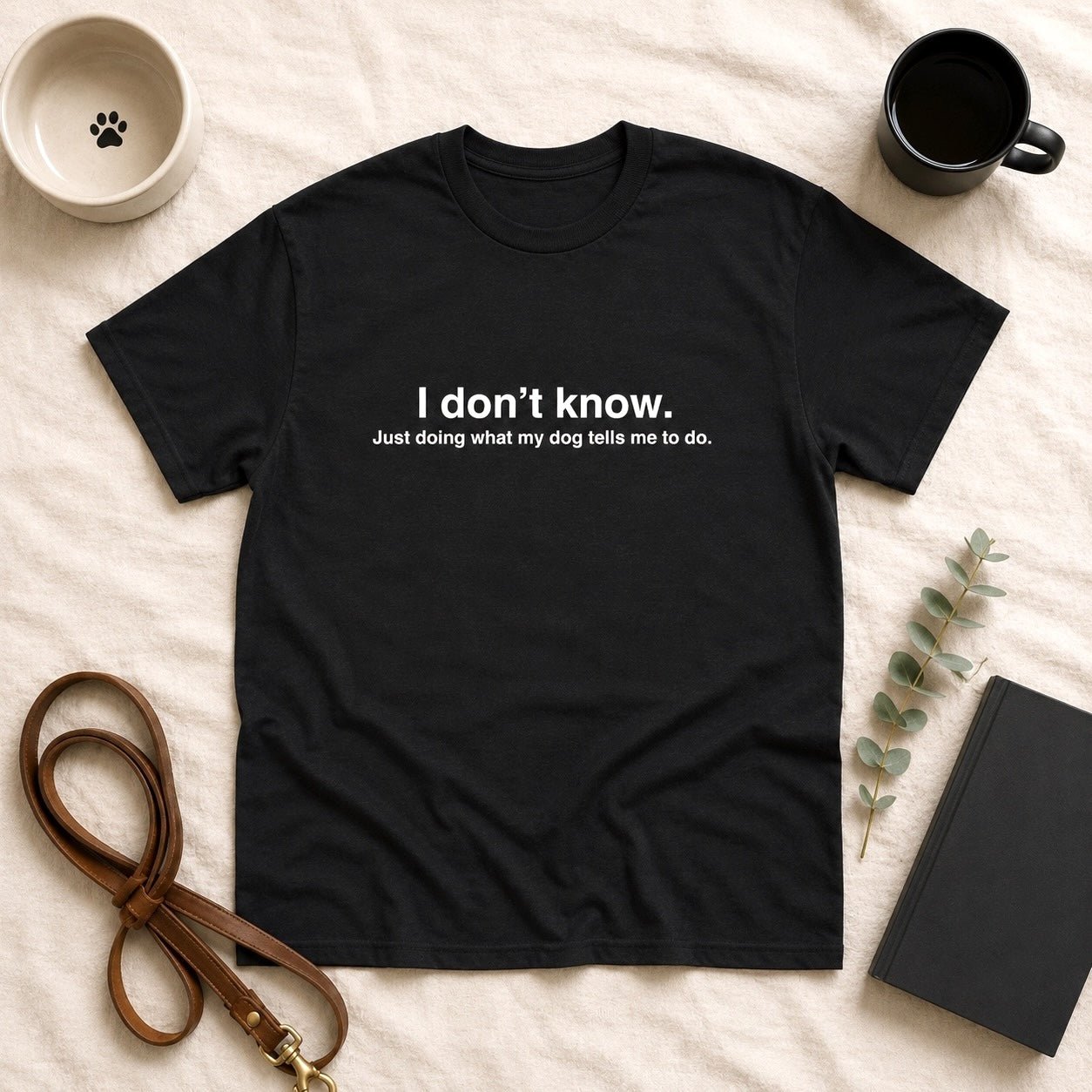Black t-shirt flat-lay with white text reading "I don't know. Just doing what my dog tells me to do." styled with a paw-print dog bowl, black coffee mug, leather leash, eucalyptus sprig, and journal on cream linen.