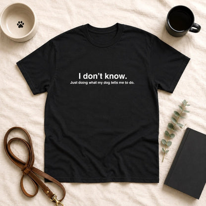 Black t-shirt flat-lay with white text reading "I don't know. Just doing what my dog tells me to do." styled with a paw-print dog bowl, black coffee mug, leather leash, eucalyptus sprig, and journal on cream linen.