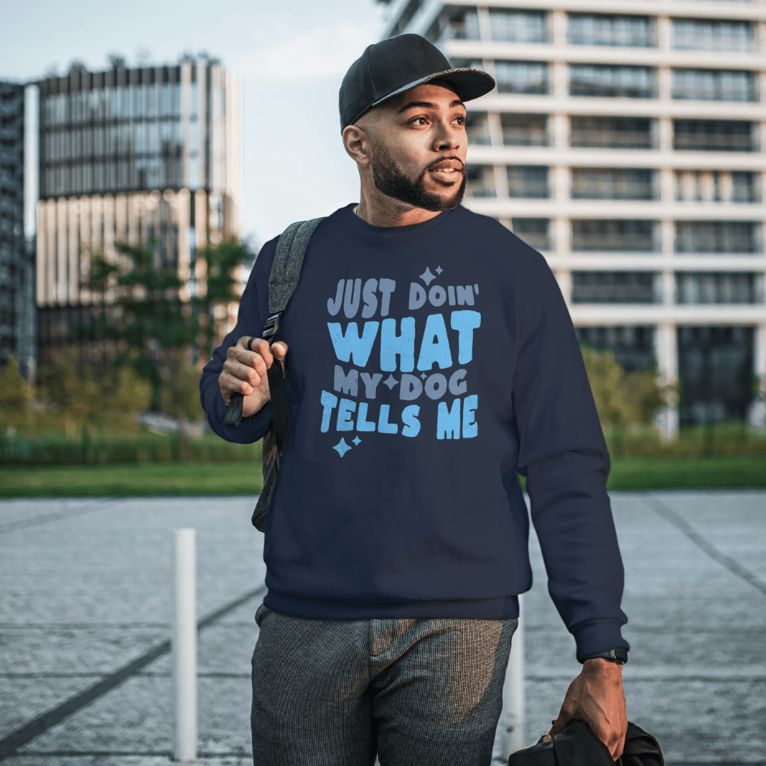 A man in a blue cap and cozy THiNK LiKE A DOG® Quirky Dog Lover Sweatshirt, featuring "Just Doing What My Dog Tells Me," strolls outdoors near modern buildings.