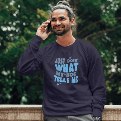 A man with a bun hairstyle, smiling and chatting on the phone, wears a dark THiNK LiKE A DOG® Quirky Dog Lover Sweatshirt – ‘Just Doing What My Dog Tells Me,’ against a cozy backdrop of trees.