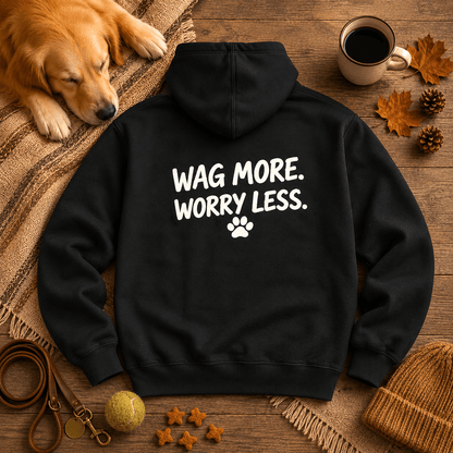 Black Wag More. Worry Less. hoodie styled flat on warm wood with a leather leash, chewed tennis ball, treats, and a coffee mug.