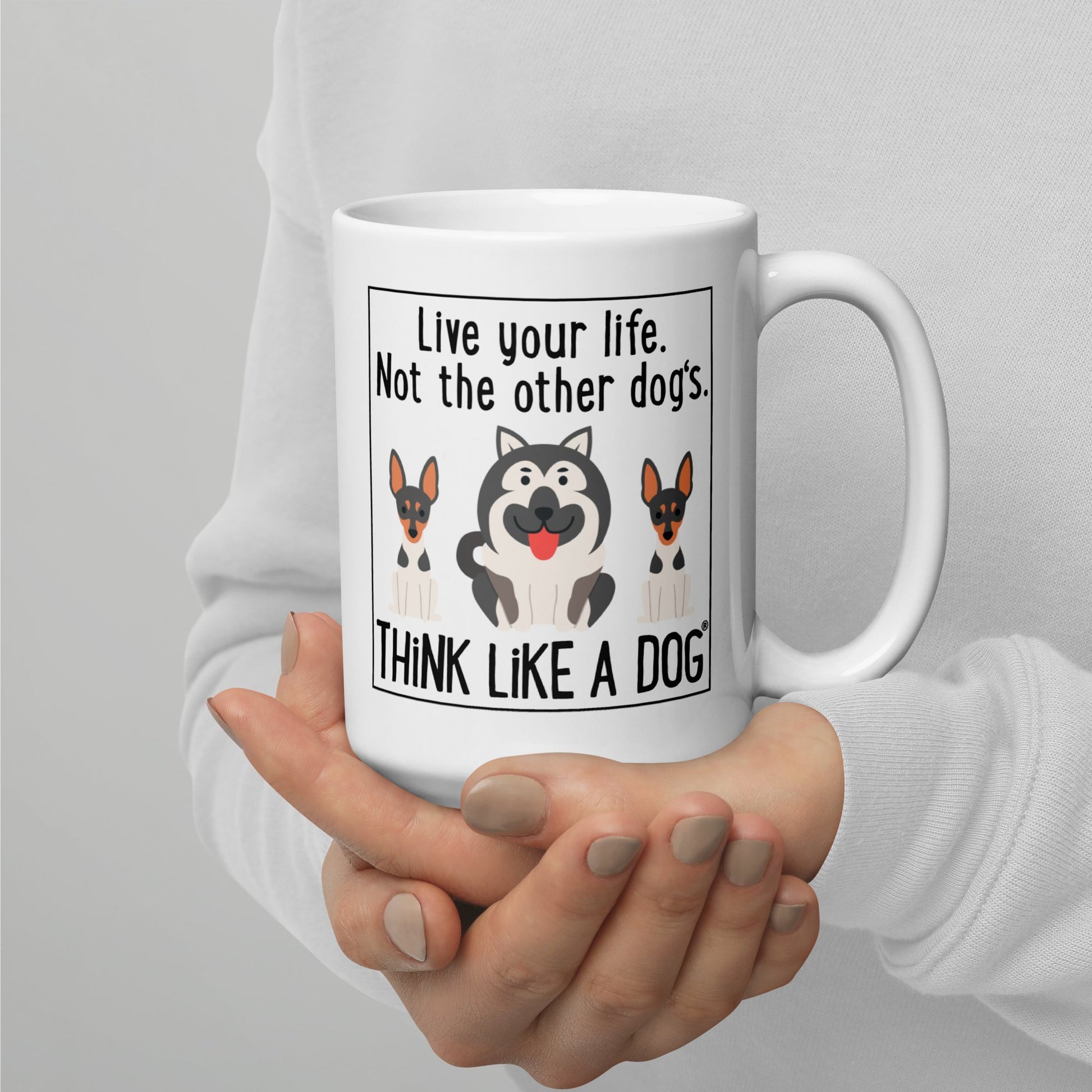 A person holding a THiNK LiKE A DOG® white glossy mug with text "live your life. not the other dog’s. think like a dog" and illustrations of three cartoon dogs.