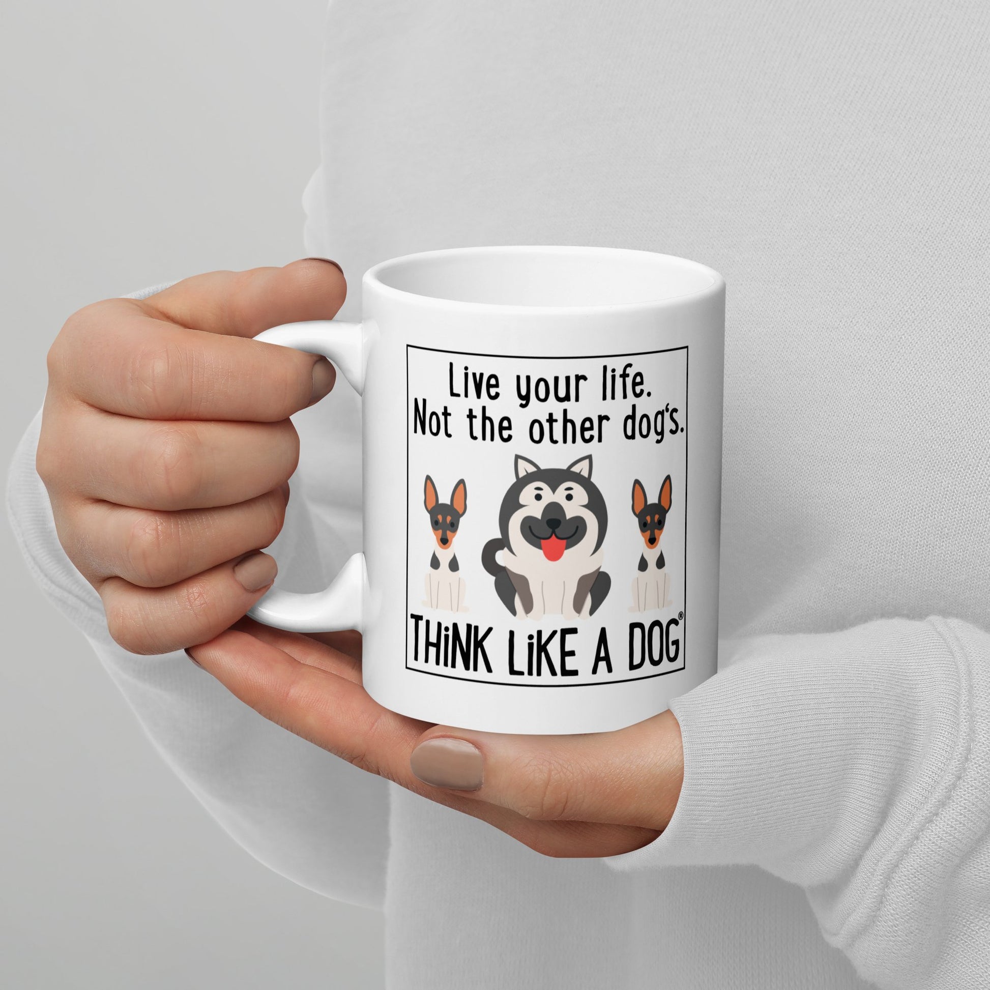 A person holding a THiNK LiKE A DOG® white glossy mug with the text "live your life, not the other dog's. think like a dog" and cartoon dogs.