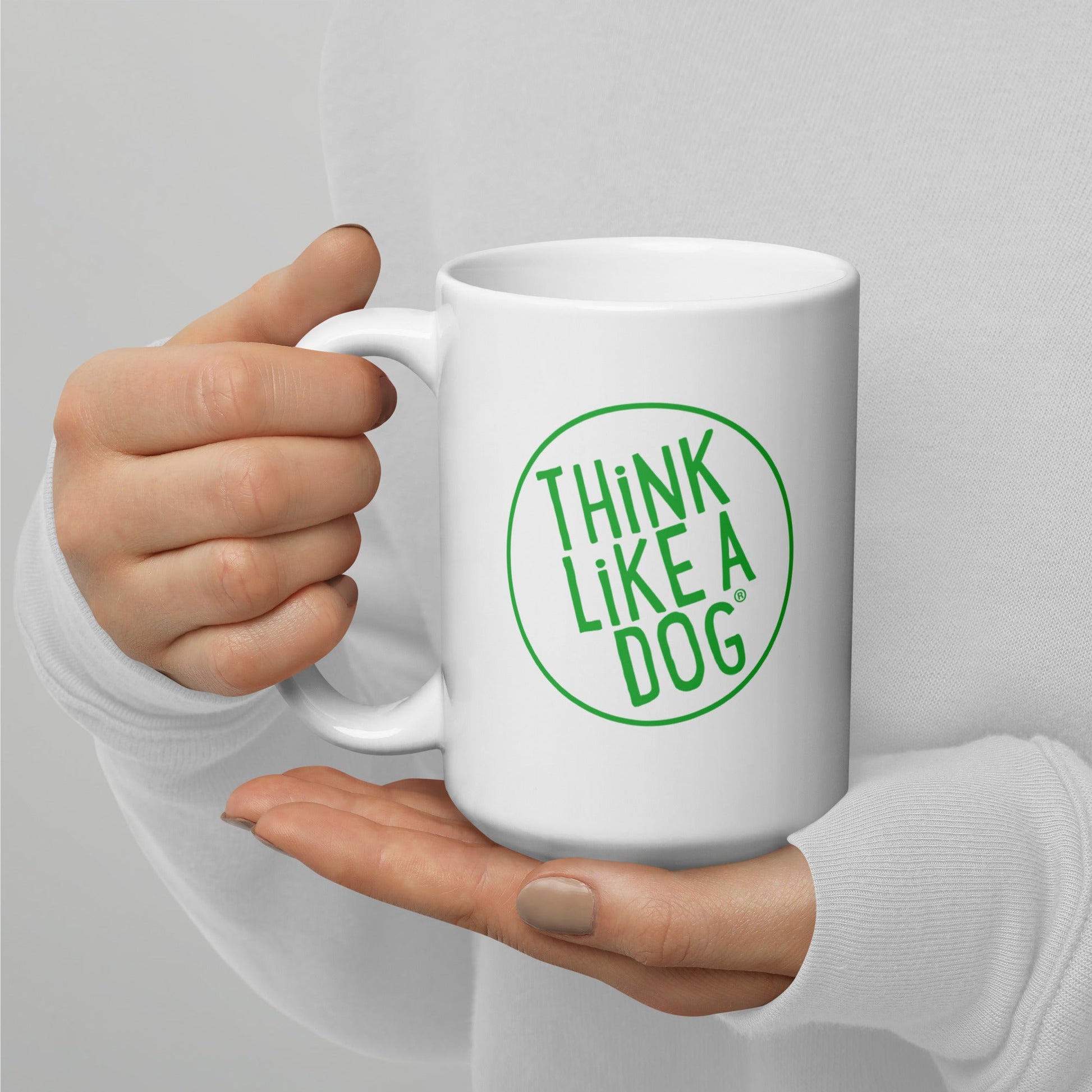 A person holding a white mug with their right hand. The mug has a text design that reads 'THINK LIKE A DOG' in bold, green letters inside a green circle outline.