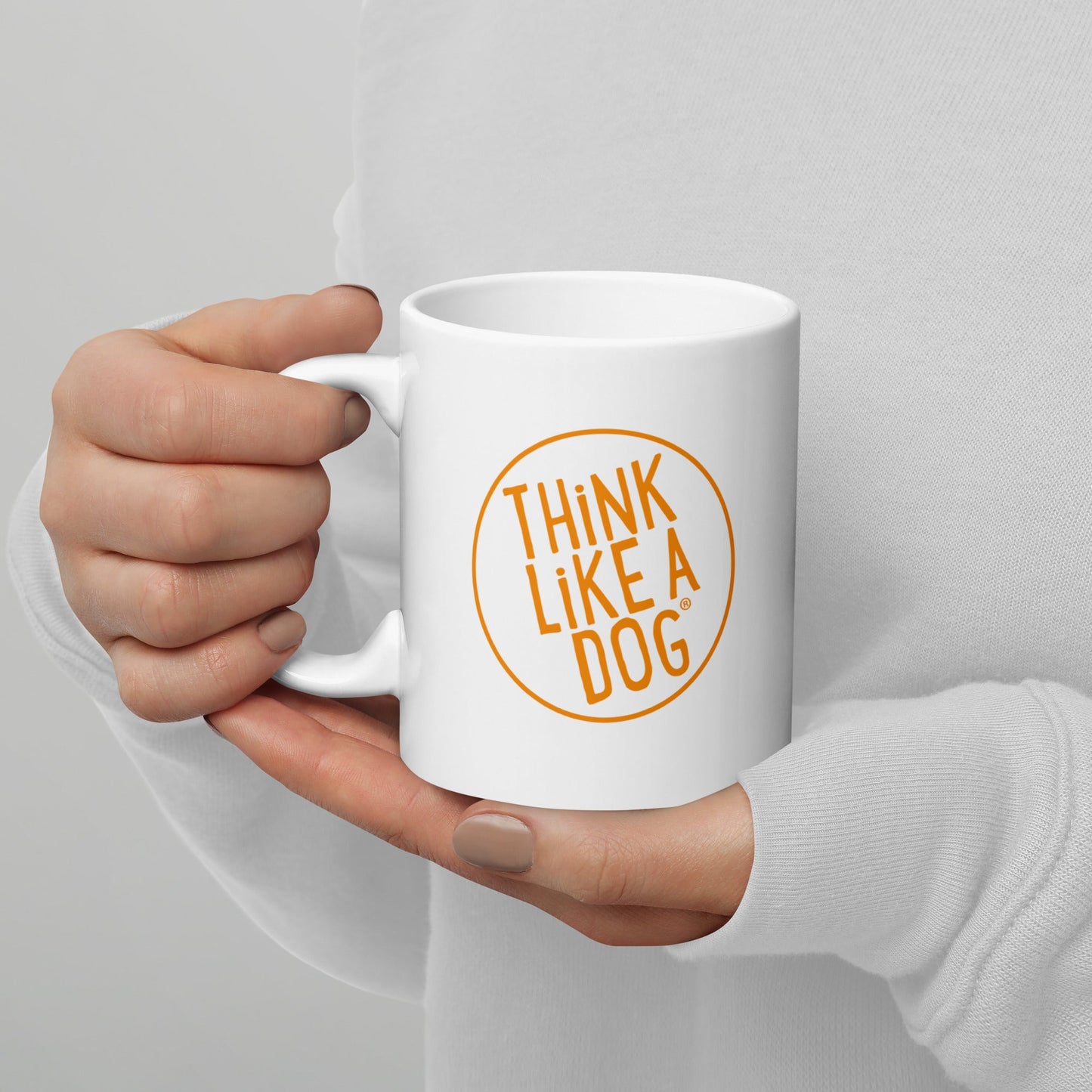 A person holding a white mug with their right hand. The mug has a text design that reads 'THINK LIKE A DOG' in bold, orange letters inside an orange circle outline.