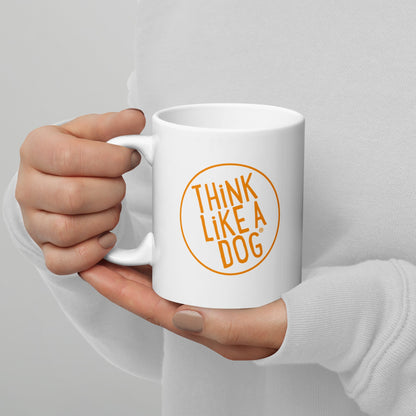 A person holding a white mug with their right hand. The mug has a text design that reads 'THINK LIKE A DOG' in bold, orange letters inside an orange circle outline.