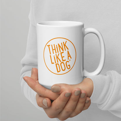 A person holding a white mug with their right hand. The mug has a text design that reads 'THINK LIKE A DOG' in bold, orange letters inside an orange circle outline.