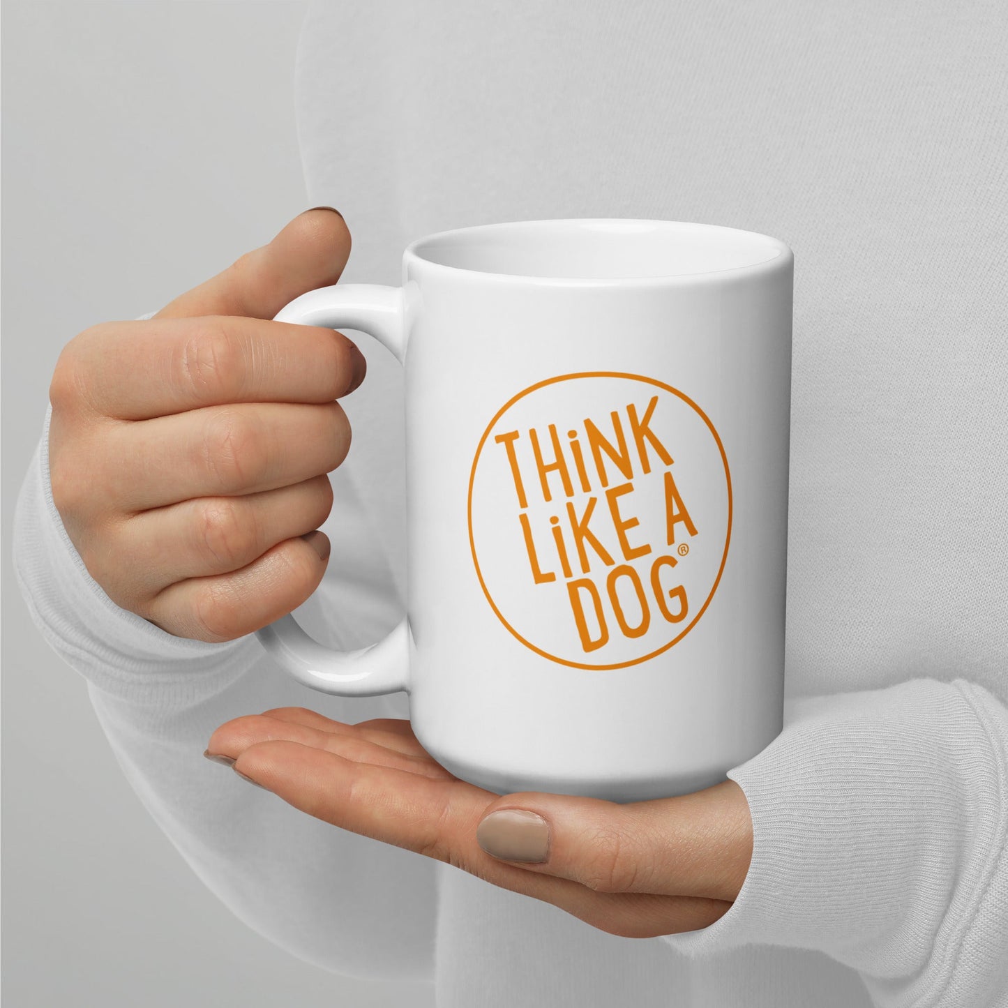 A person holding a white mug with their right hand. The mug has a text design that reads 'THINK LIKE A DOG' in bold, orange letters inside an orange circle outline.