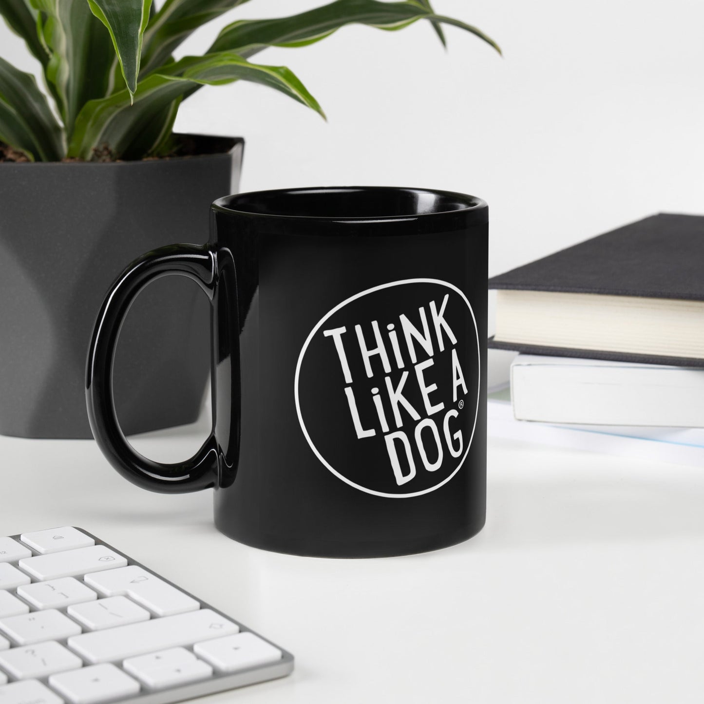 A black glossy mug with a handle on the left, featuring the white text 'THINK LIKE A DOG' inside a white circle outline. The mug is on a desk next to a plant in a black pot, a keyboard, and stacked books.